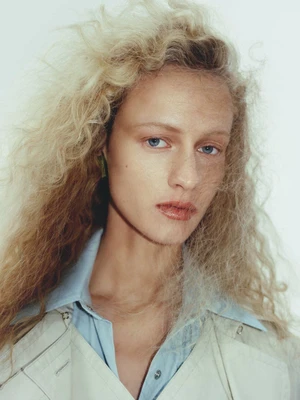 Close-up portrait of a woman with blonde curly hair wearing a light-colored coat and blue collared shirt against a white backdrop.