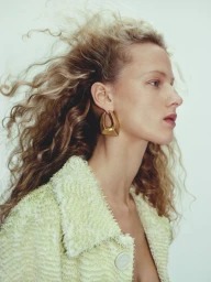 Profile portrait of a blonde woman with curly hair wearing a textured light green jacket and chunky gold earrings against a light background.