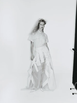 Model with voluminous curly hair wearing a button-down shirt and long sheer layered skirt against a white studio background.