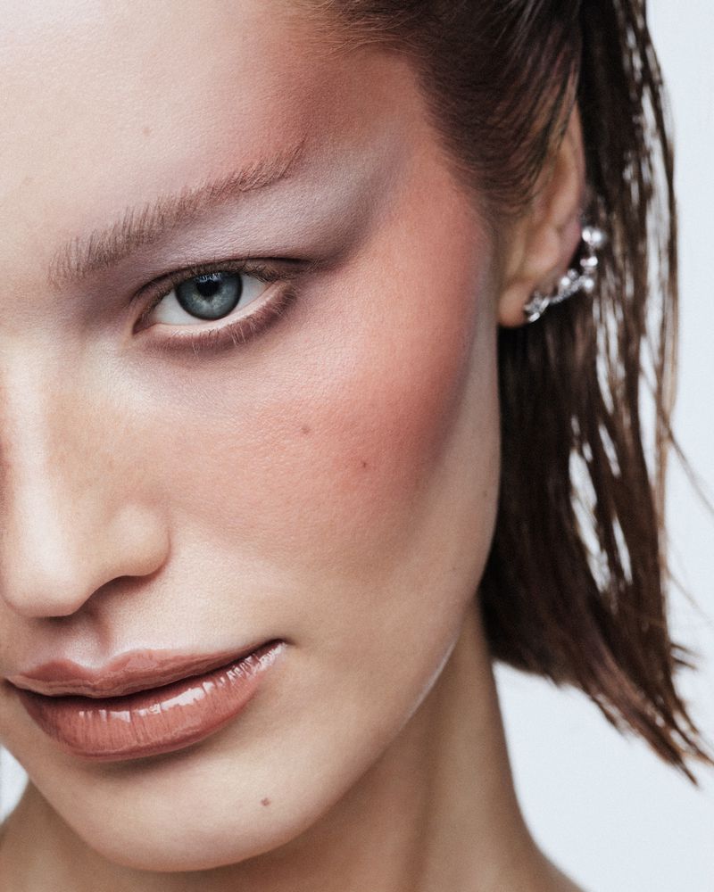 Close-up studio shot of a young adult woman with distinctive makeup including prominent blush and eyeshadow, and wet-look hair.