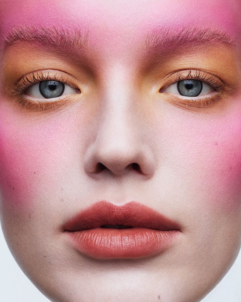 Close-up portrait of a fair-skinned person with pink and gold makeup, light blue eyes, and a neutral facial expression on a neutral background.