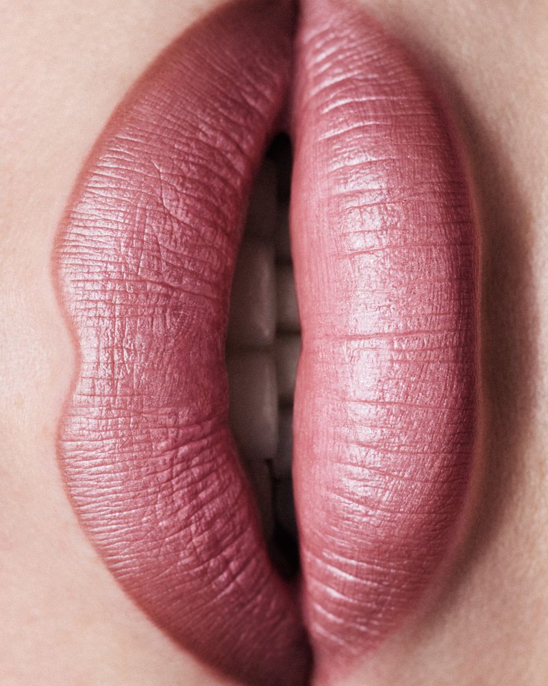Close-up of woman's lips with mauve lipstick and visible teeth. Beauty shot emphasizing the texture and color of the makeup.