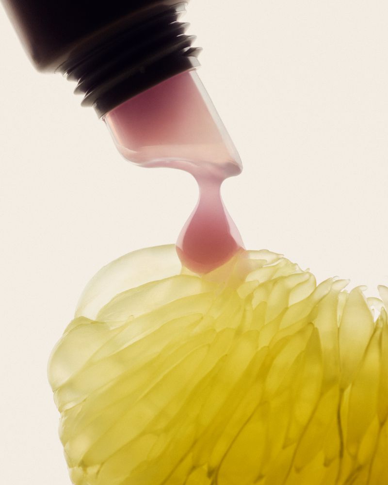 Close-up studio shot of liquid from a cosmetic tube dripping onto a semi-translucent grapefruit segment on a white surface.