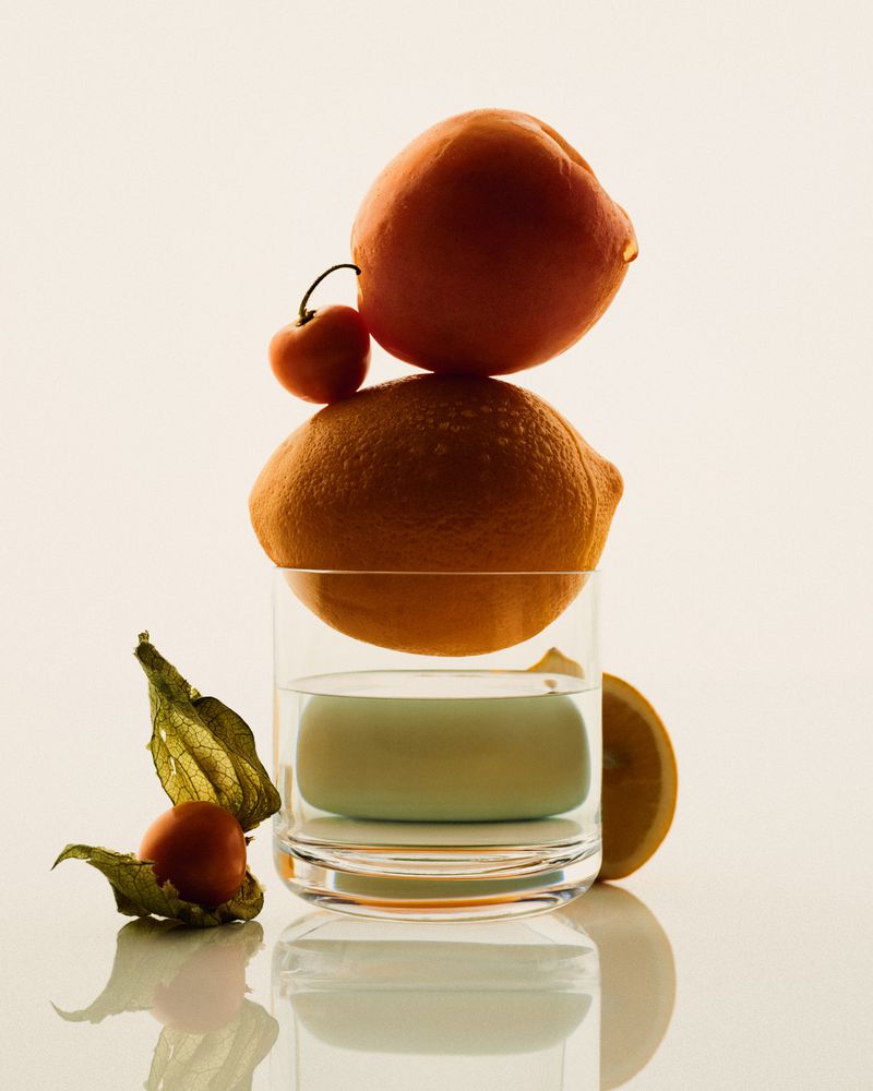 Balanced stack of citrus fruit and a cherry on a water glass against a pale background