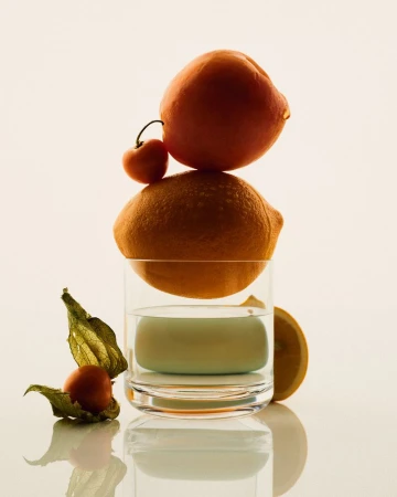 Balanced stack of citrus fruit and a cherry on a water glass against a pale background