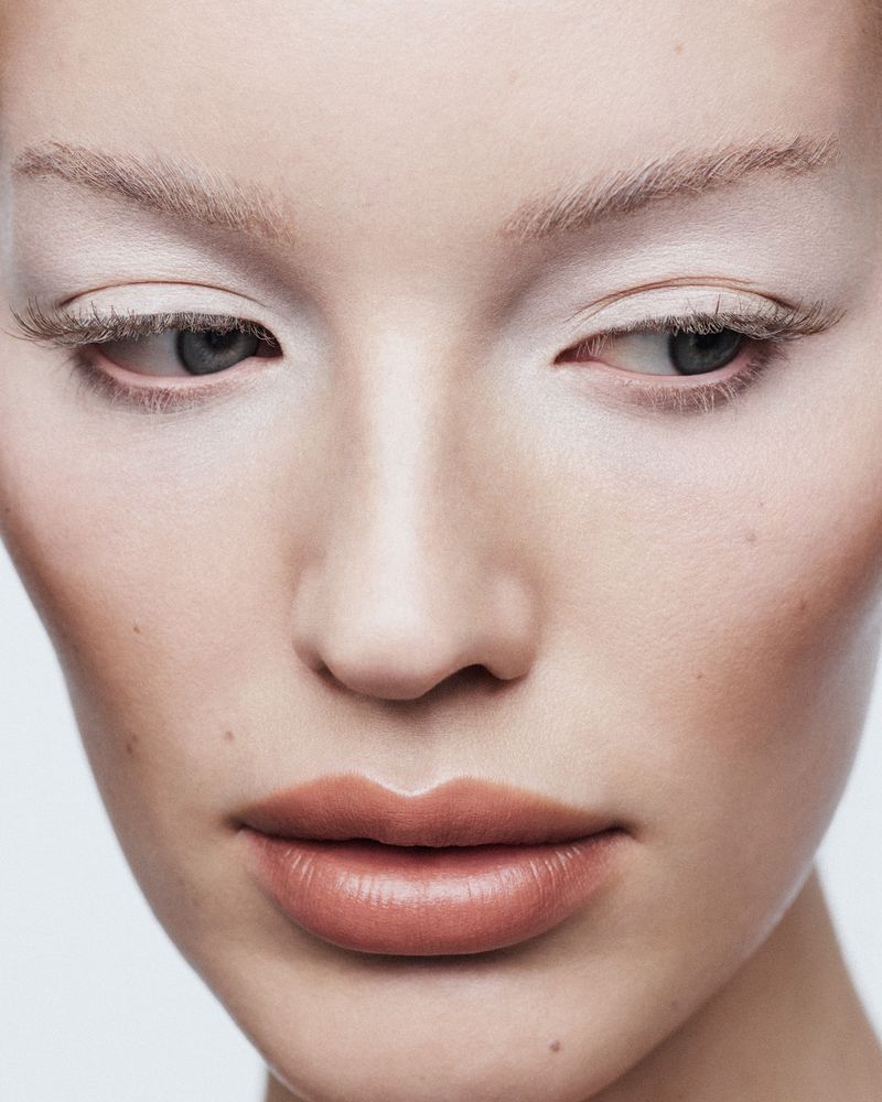 Close-up of an adult female face with cream-colored makeup, visible face and shoulder against a bright background in a beauty setting.