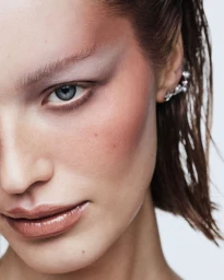 Close-up portrait of a woman with wet hair, pale pink eyeshadow, and a pearl ear cuff against a plain white background.