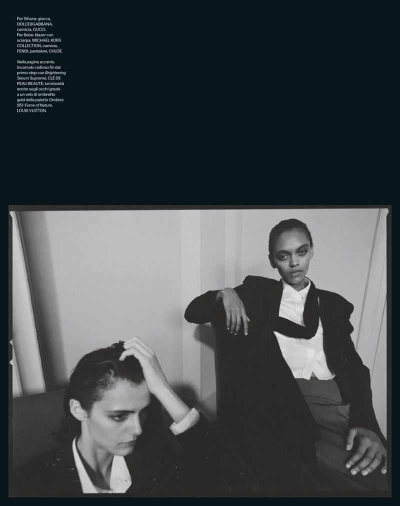 Black and white fashion shot of two models in blazers posing indoors against a wall with natural lighting, showcasing style and elegance.