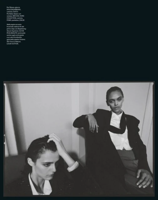 Black and white fashion shot of two models in blazers posing indoors against a wall with natural lighting, showcasing style and elegance.
