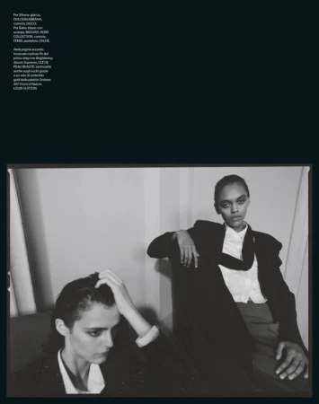 Black and white fashion shot of two models in blazers posing indoors against a wall with natural lighting, showcasing style and elegance.