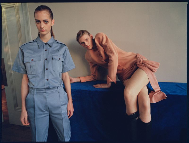 Two young women in a photo studio fashion shoot. One stands in a pale blue suit; the other reclines in a light peach jacket dress.