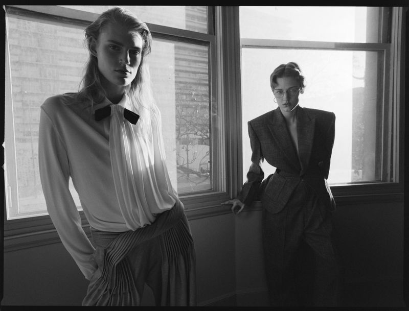 Two people in an indoor setting. One person wearing a white shirt and pleated pants; the other wearing a grey suit. Window light.