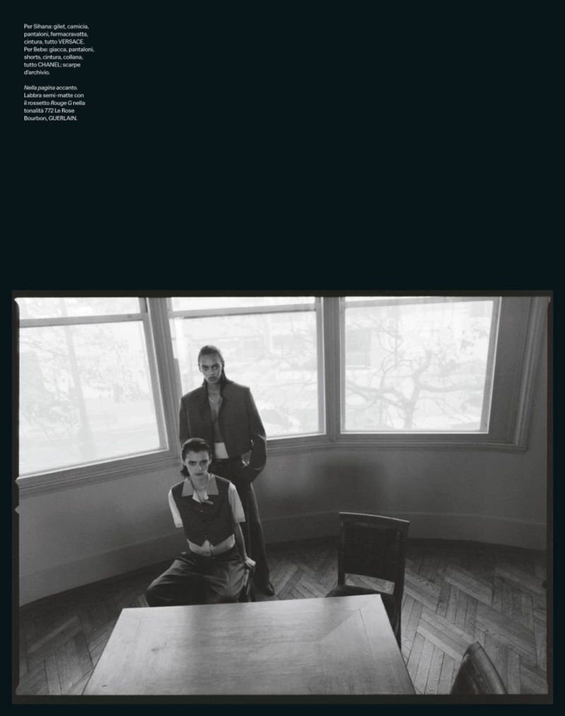 Two people in black and white fashion photo. One adult is seated at a table, the other standing behind near a window in a room.