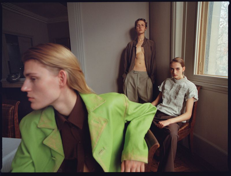 Three fashion models pose indoors wearing casual jackets and pants; one model is seated by a window with natural light.