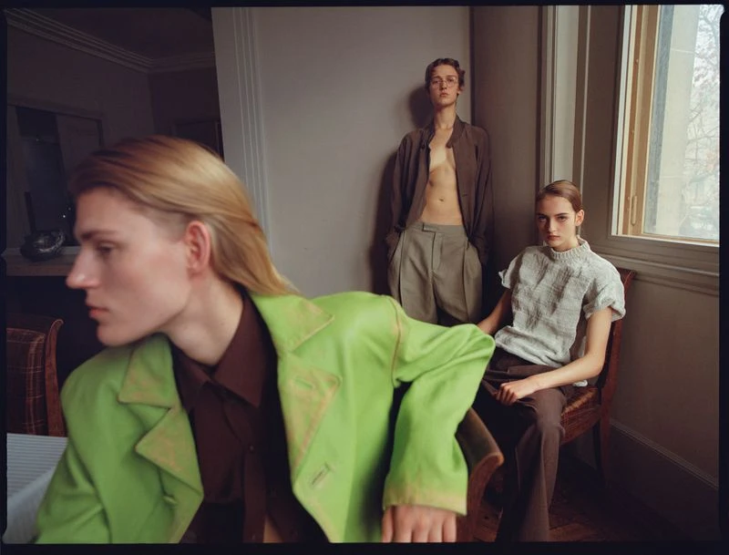 Three fashion models pose indoors wearing casual jackets and pants; one model is seated by a window with natural light.