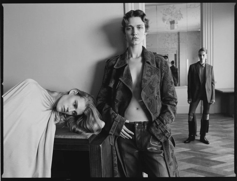 Black and white fashion shot of three women in an indoor setting, showcasing stylish outerwear and dramatic poses in a minimalist scene.
