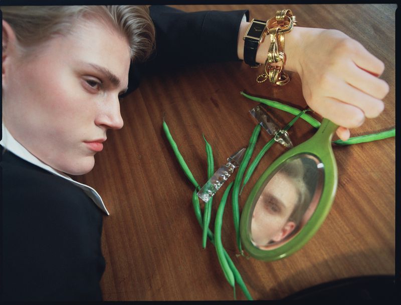 Blond male model in suit poses on a table with a hand mirror, green beans, a watch, and jewelry in a fashion editorial shot.