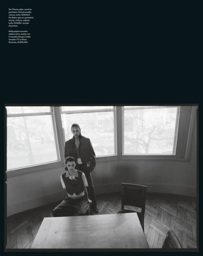 Black and white editorial image of two models in a room with a large bay window and wooden table.