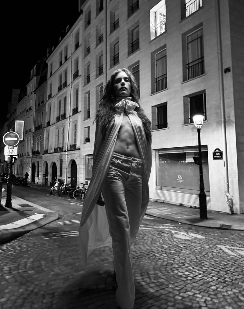 Black and white shot of a woman in white pants and a top walking down a cobblestone street in front of Parisian buildings at night.