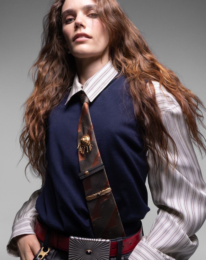 A young woman with long brown hair wears a navy vest over a striped shirt, accessorized with a decorative tie and a statement belt buckle.