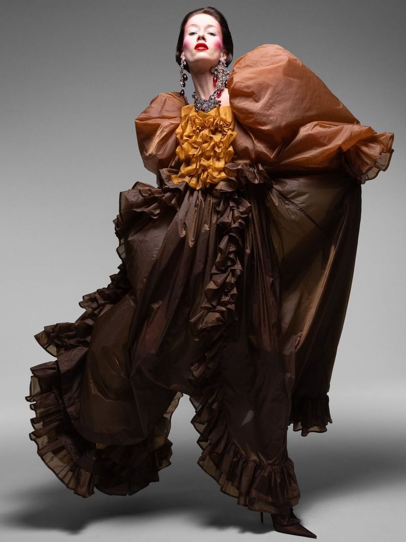 Elegant woman in sculptural brown ruffled gown with bold makeup and jewelry strikes a pose in a studio fashion editorial.