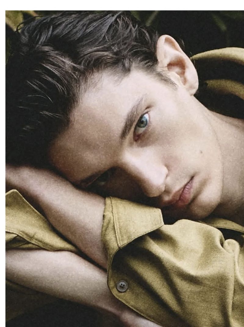 Close-up portrait of a handsome young adult male with dark hair and blue eyes, wearing a yellow collared shirt, resting his head on arms.