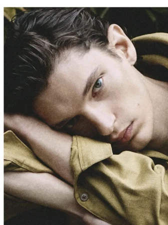 Close-up portrait of a handsome young adult male with dark hair and blue eyes, wearing a yellow collared shirt, resting his head on arms.