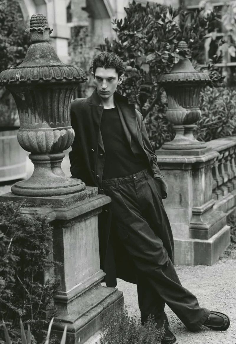 Monochrome portrait of a slim adult male leaning against a stone wall in an outdoor park-like setting with foliage in the background.