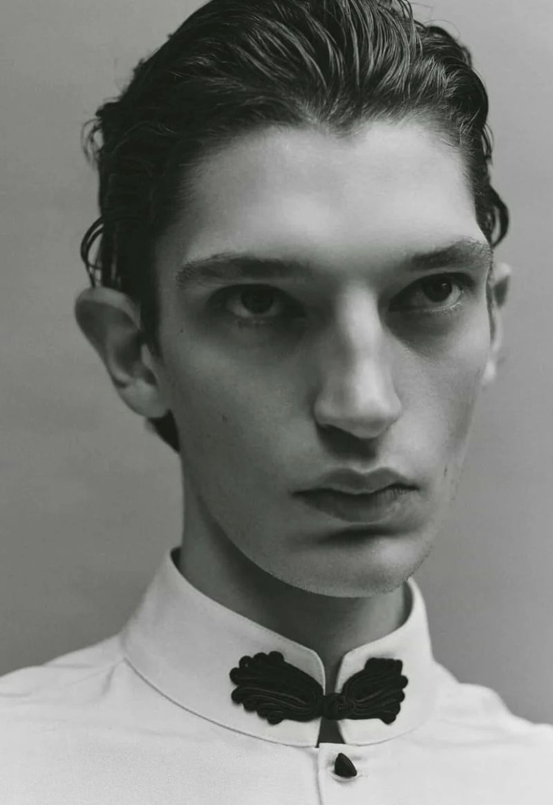 Striking close-up of a young adult male with dark hair in a white shirt with a decorative black collar, intense gaze, monochrome.