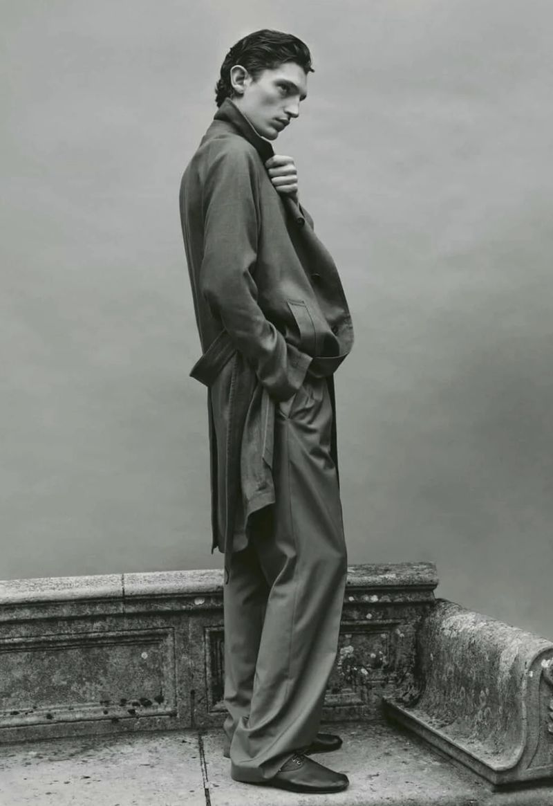 A man in a suit stands in front of a plain backdrop, posing on a stone ledge, wearing a loose jacket and trousers with loafers.