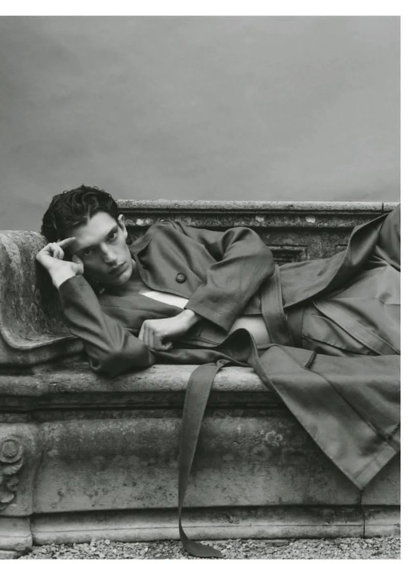 Adult male model lying on a stone ledge wearing a suit, set against a grayscale background, capturing a contemplative mood.