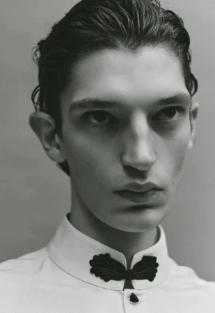 Close-up portrait of a male model in a white shirt with black decorative collar embroidery against a neutral studio backdrop