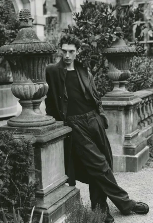 Monochrome editorial portrait of a man leaning against a stone balustrade with decorative urns outdoors