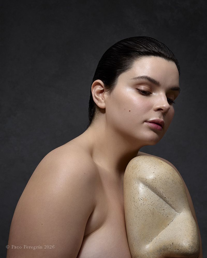 Nude woman embraces a light-tan abstract sculpture with her eyes closed in a studio portrait, illuminated by soft, natural light.