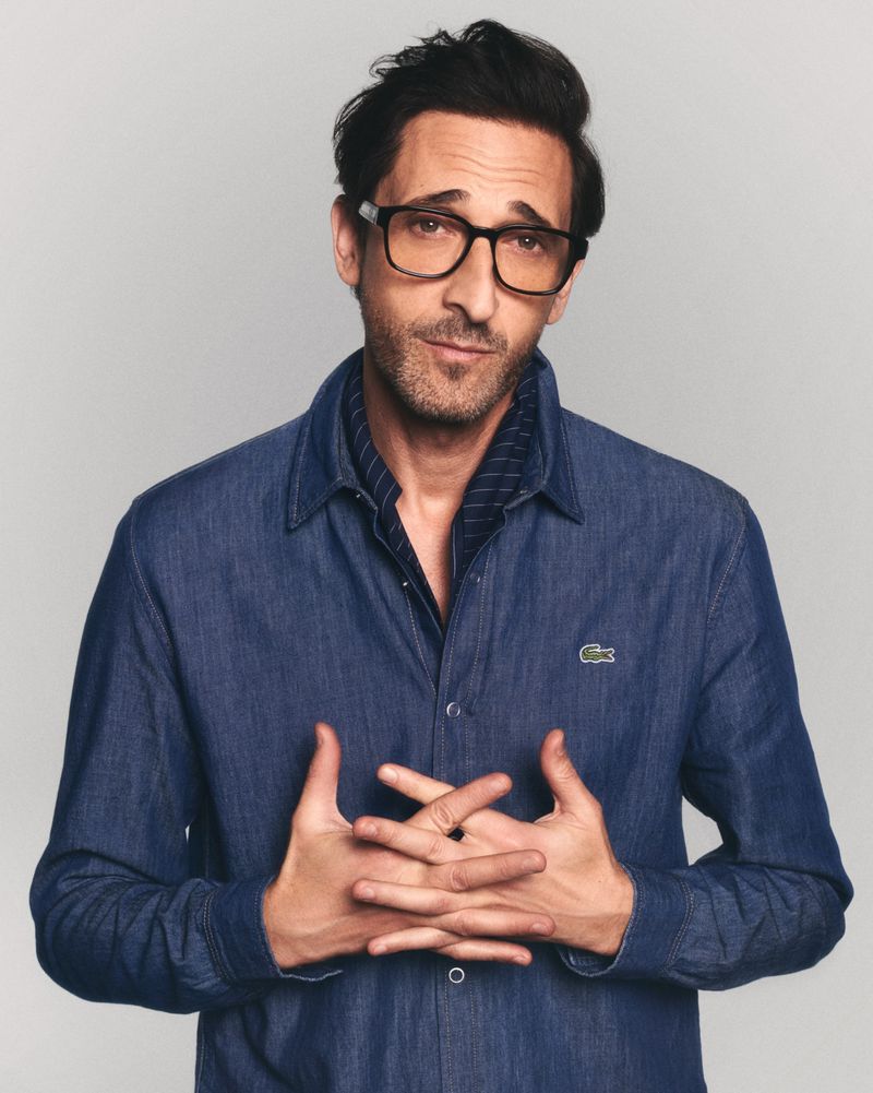 Portrait of a man wearing glasses and a blue denim shirt. He is looking at the camera with a slight smile, hands clasped in front.
