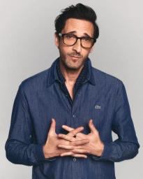 Adrien Brody wearing glasses and a denim shirt against a plain light grey backdrop for Lacoste Eyewear