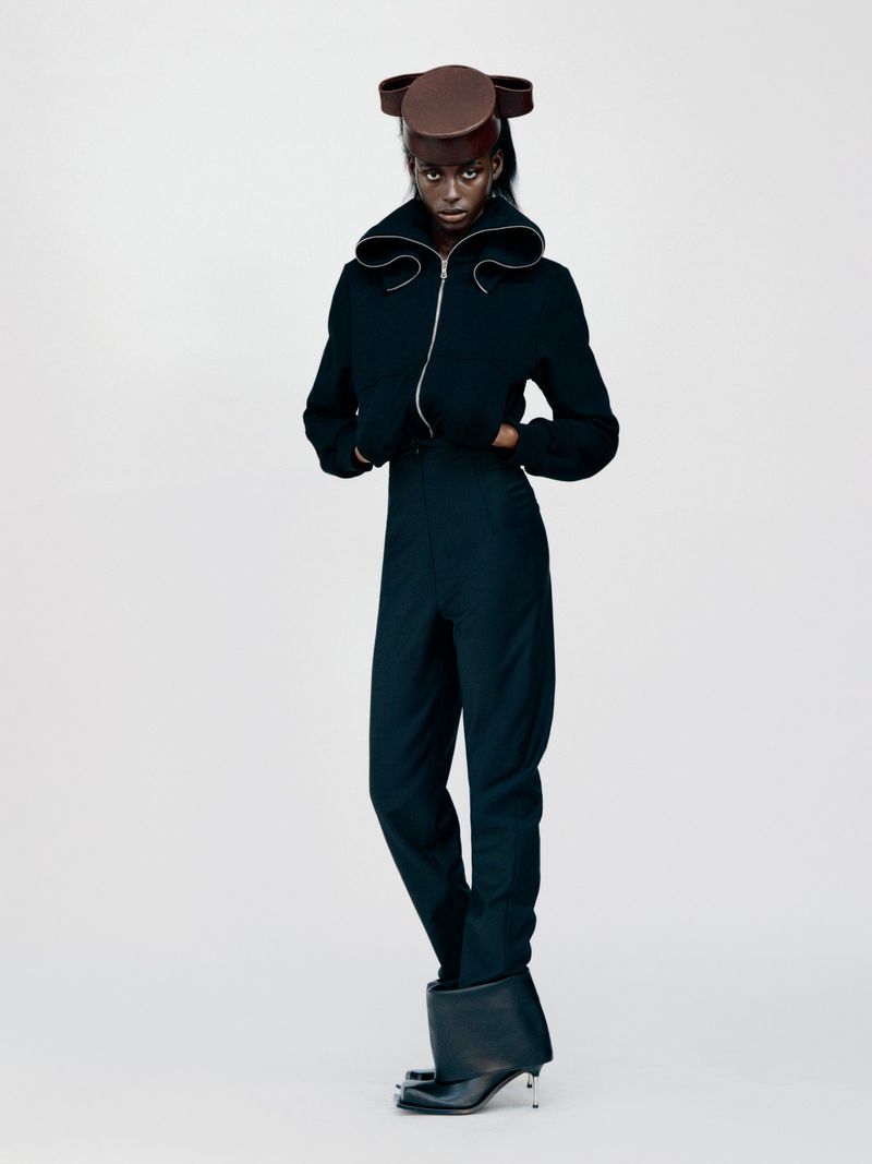 Full-length studio shot of a woman with dark skin wearing a dark jumpsuit, jacket, hat, and heeled boots against a white backdrop.