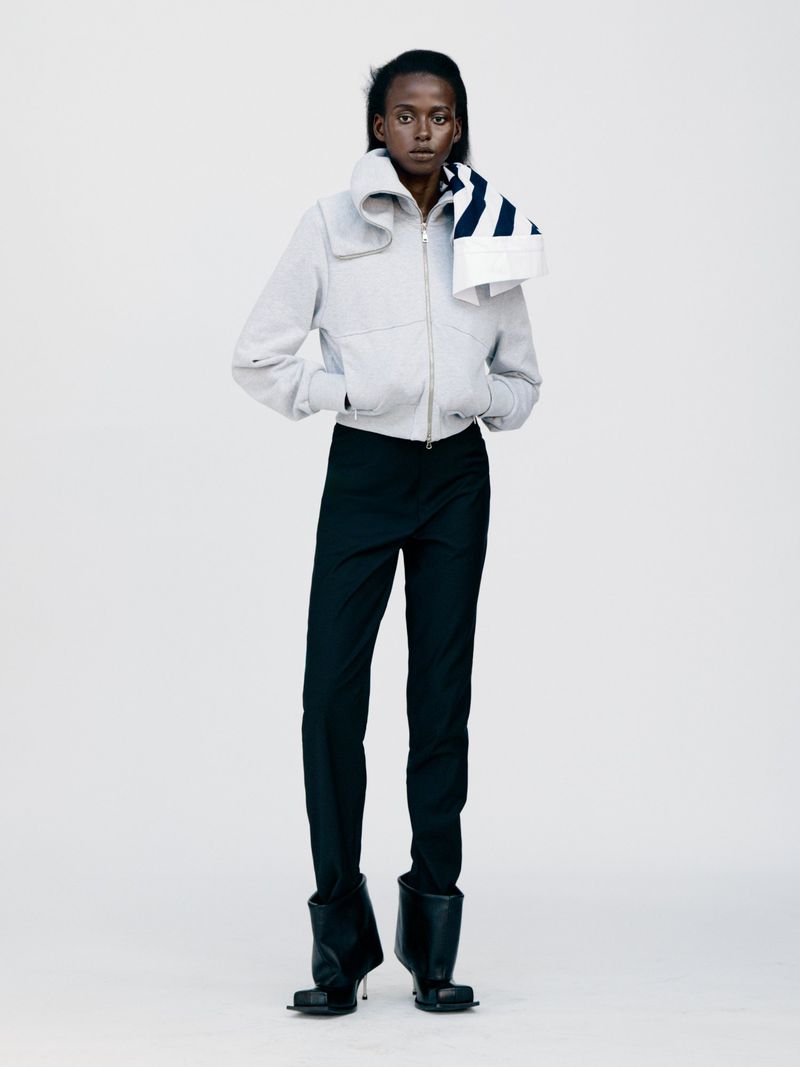 Fashionable woman in studio setting wearing light jacket, black pants, and sculptural black boots, posing in a modern, editorial style.