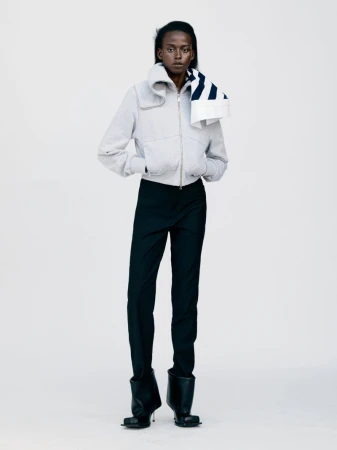 Fashionable woman in studio setting wearing light jacket, black pants, and sculptural black boots, posing in a modern, editorial style.