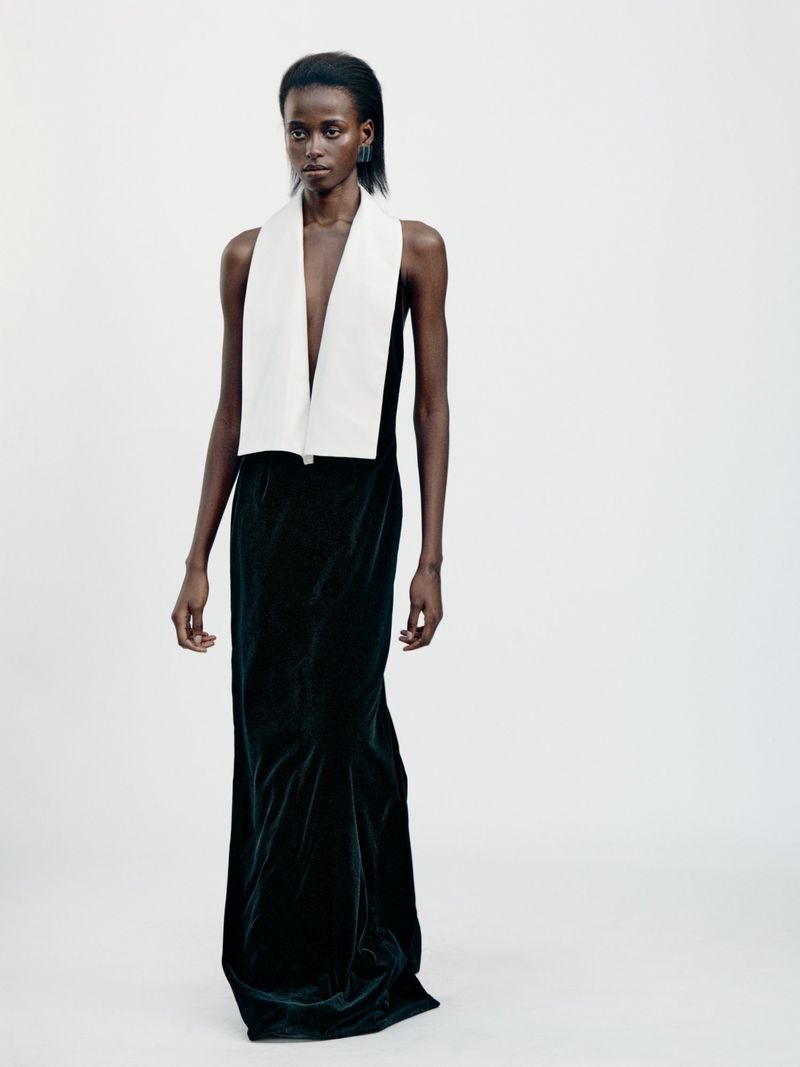 Elegant black female model poses in a long, dark dress with a contrasting white stole in a brightly lit studio setting.