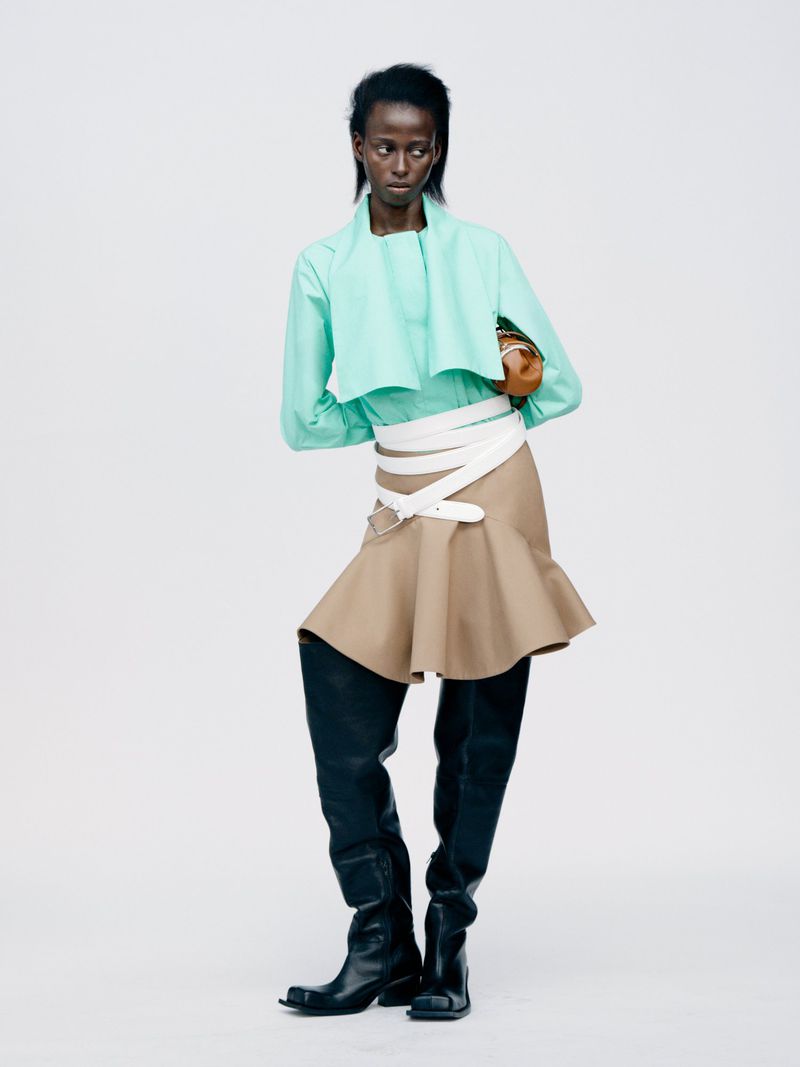 Fashion shot of a Black woman in a mint green top, tan skirt, and black leather boots against a white background in a studio setting.