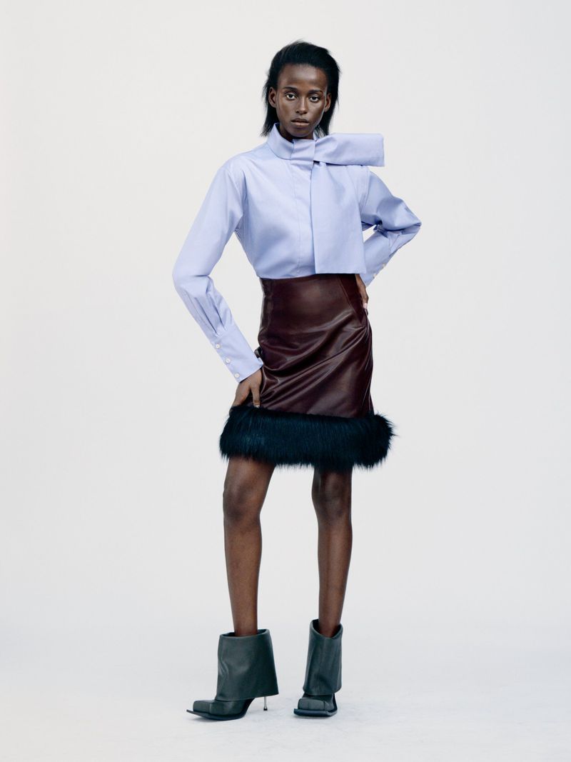Model wearing a light blue bow-collar shirt, dark leather skirt with faux fur trim, and black boots against a white backdrop