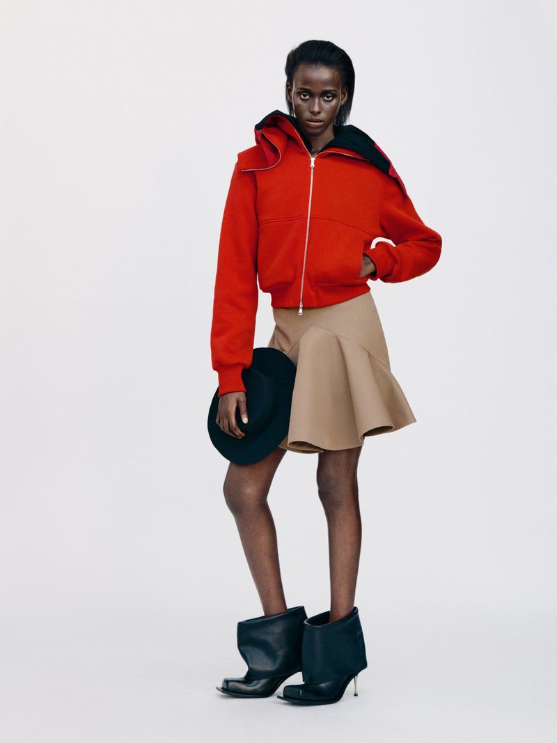 Full-length fashion shot of a solo adult female wearing a red hooded jacket, tan skirt, and black heeled boots in a white studio.