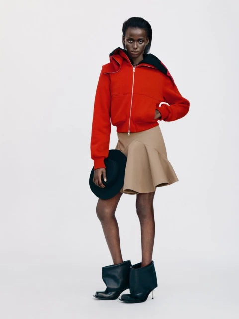Full-length fashion shot of a solo adult female wearing a red hooded jacket, tan skirt, and black heeled boots in a white studio.