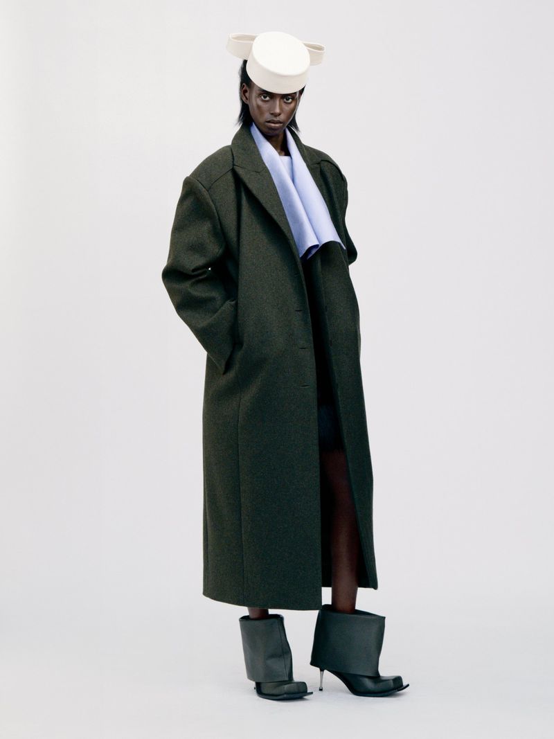 A tall, thin Black woman stands in a photo studio wearing an overcoat, light-blue scarf, hat and dark boots against a white backdrop.