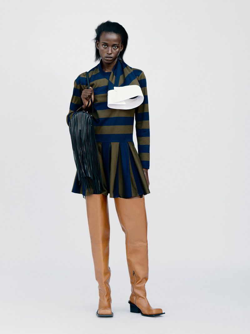 Full-length fashion shot: a Black female model poses in a striped dress with thigh-high boots and a fringe bag on a plain background.