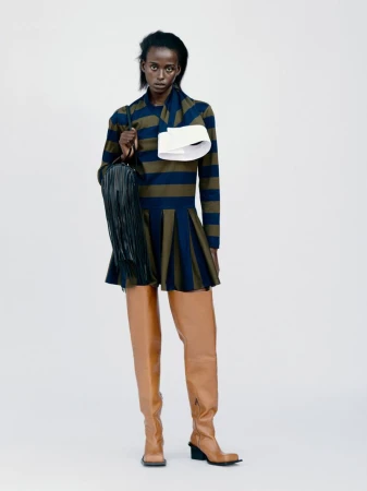 Full-length fashion shot: a Black female model poses in a striped dress with thigh-high boots and a fringe bag on a plain background.