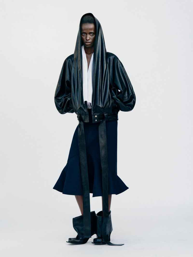 Editorial fashion shot of a black female model wearing a black bomber jacket, navy skirt, and black leather boots in a bright photo studio.
