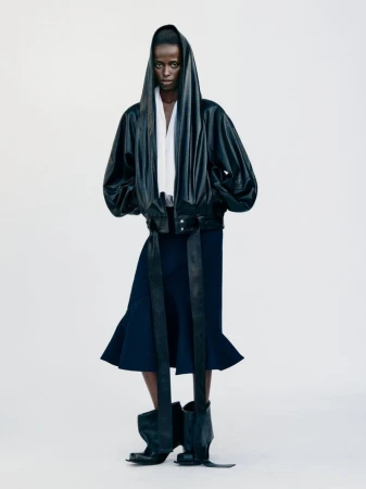 Editorial fashion shot of a black female model wearing a black bomber jacket, navy skirt, and black leather boots in a bright photo studio.