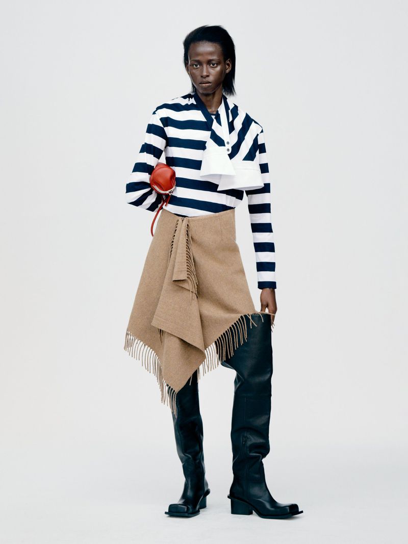 An adult woman in a blue and white striped long sleeve shirt, tan skirt, and black boots poses in front of a white background.